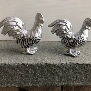 May include: Two silver rooster figurines with black detailing. The roosters are facing different directions and have their tails raised.