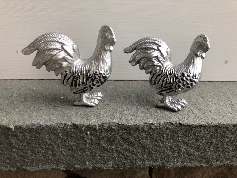 May include: Two silver rooster figurines with black detailing. The roosters are facing different directions and have their tails raised.