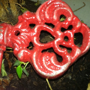 May include: A red metal key-shaped ornament with intricate details. The ornament has a shiny, glossy finish and is surrounded by dirt and foliage.