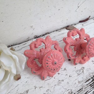 May include: Two pink metal decorative hooks with ornate designs. The hooks have a distressed finish and are mounted on a white painted surface.