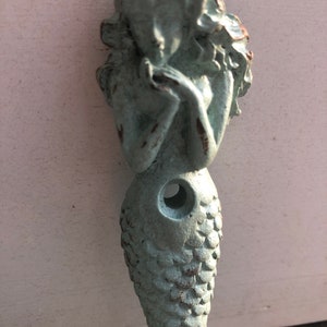 May include: A light blue mermaid figurine with a scaly tail and three holes for hanging. The mermaid is holding her hands together in front of her face.