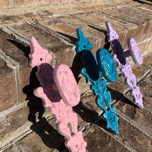 May include: Three ornate door knobs in pink, teal, and lavender. The knobs are made of metal and have a decorative design. They are arranged in a row on a brick surface.