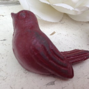 May include: A small, red, cast iron bird figurine. The bird is perched on a white surface.