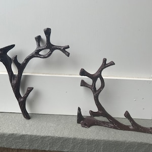 May include: Two cast iron wall hooks shaped like branches with intricate details. The hooks are painted a dark brown color and have a rustic look.