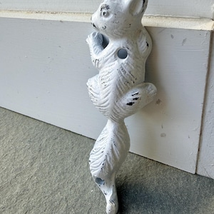 May include: A white cast iron squirrel door knocker. The squirrel is in a climbing pose with its tail extending downward. The door knocker has a distressed finish with black accents. It is mounted on a white door frame.