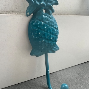 May include: A teal blue pineapple shaped cast iron hook. The hook is designed to look like a pineapple with a leafy top and a textured body. The hook is attached to a wall.