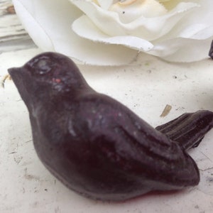 May include: A dark purple bird ornament with a smooth, glossy finish. The bird is in a perched position, with its tail feathers slightly raised. A white flower and a weathered, white surface are in the background.