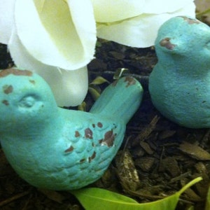 May include: Two teal blue bird figurines with distressed paint, sitting on a bed of brown mulch.