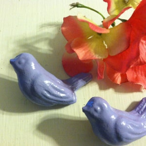 May include: Two small, lavender blue ceramic birds with a glossy finish. The birds are perched on a white surface.