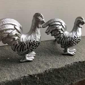 May include: Two silver rooster figurines with detailed feathers and textured surfaces.
