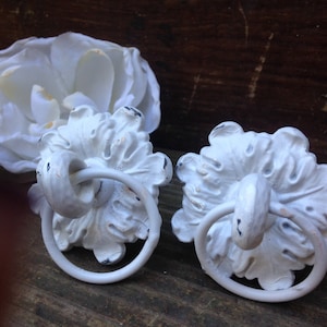 May include: Two white, ornate, decorative drawer pulls with a ring handle. The pulls have a floral design and are made of metal.
