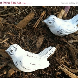 May include: Two white painted metal bird figurines with black eyes. The birds are sitting on a bed of brown wood chips.