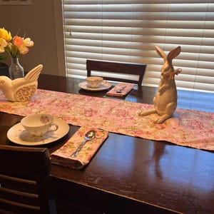 May include: A wooden table set with a pink floral table runner, a white ceramic bird figurine, a white ceramic rabbit figurine, a white teacup and saucer, a silver spoon, and a floral napkin.