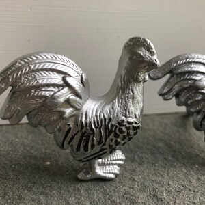 May include: Two silver rooster figurines with black accents. The roosters are standing on a grey surface.