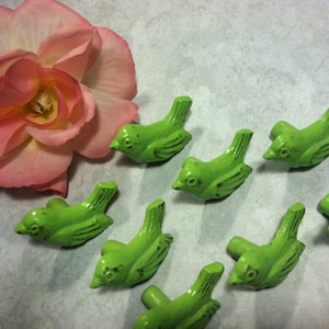 May include: Eight green bird-shaped cabinet knobs with a distressed finish. The knobs are arranged in a semi-circle on a light grey surface.