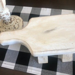 May include: White wooden stool with a distressed finish. The stool has a rectangular top and a single handle on the side. The stool is sitting on a black and white checkered surface.