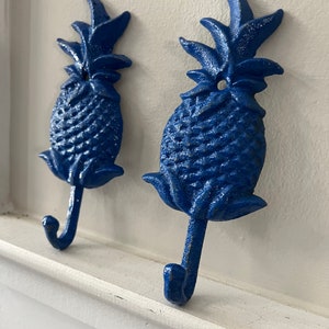 May include: Two blue cast iron pineapple shaped hooks mounted on a white wall.