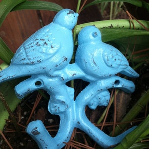 May include: Two blue bird-shaped hooks are attached to a branch. The hooks are made of metal and have a distressed finish. The branch is also blue and has a distressed finish.