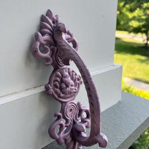 May include: A decorative, ornate, lavender-colored door pull. The metal hardware features a detailed design with a curved handle and a central medallion with a face. The pull is mounted on a white surface.