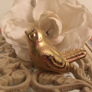 May include: A gold-painted bird figurine with a detailed design, perched on a white decorative surface.