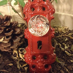 Peut inclure: Une plaque de porte décorative rouge avec un bouton en verre transparent. La plaque a un design orné et un trou de serrure. Le bouton est rond avec un motif tourbillonnant. La plaque de porte est placée dans une jardinière avec une pomme de pin et une plante.