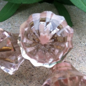 May include: Three pink glass cabinet knobs with a faceted design. The knobs have a brass screw on the back.