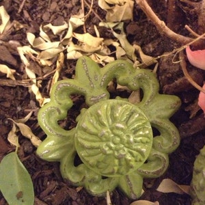 May include: A green metal door knob with a floral design. The knob is surrounded by brown mulch and dried leaves.