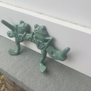 May include: A pair of green frog hooks mounted on a white wall. The frogs are perched on a branch with their legs forming hooks.
