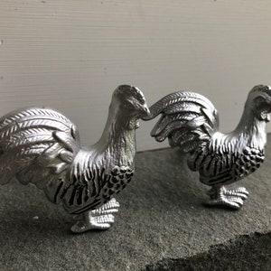 May include: Two silver rooster figurines with detailed feathers and black accents. The roosters are facing each other with their heads turned slightly.