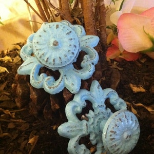 May include: Two blue metal decorative drawer pulls with ornate designs. The pulls are on a bed of brown mulch.