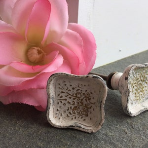 May include: Two white ceramic cabinet knobs with a gold floral design. The knobs are square shaped with rounded edges.