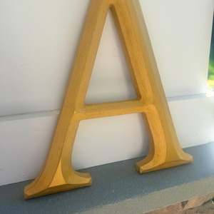 May include: A three-dimensional capital letter A finished in a metallic gold paint. The letter features a beveled edge design with a smooth, consistent surface texture and a wide base, standing upright with a clear, sharp geometric form.