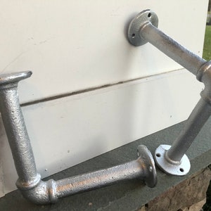 May include: Two silver metal pipe brackets with flanges. The brackets are in the shape of an L and are attached to a white wall.