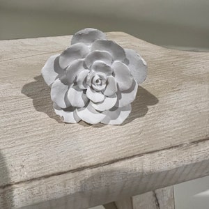 May include: A white ceramic flower with a detailed design. The flower is sitting on a white wooden surface.