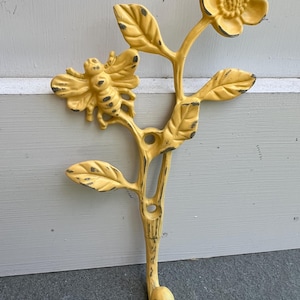 May include: Yellow metal wall hook featuring a bee and flower design. The hook is shaped like a stem with leaves and a flower at the top. The bee is positioned on the left side of the stem. The hook is painted yellow with distressed black accents.