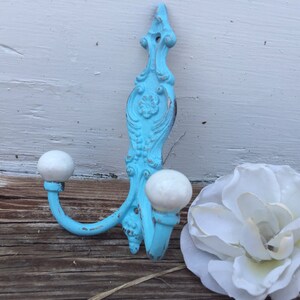 May include: A distressed, ornate, turquoise wall hook with two white ball-shaped knobs. The hook has a decorative floral design and is placed on a weathered wooden surface next to a white flower.