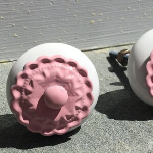 May include: Two white ceramic knobs with pink floral designs. The knobs have a raised, scalloped edge around the floral design.