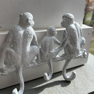 May include: Three white monkey-shaped hooks mounted on a white surface. The hooks are in a row and are facing different directions. The hooks are made of metal and have a distressed finish.