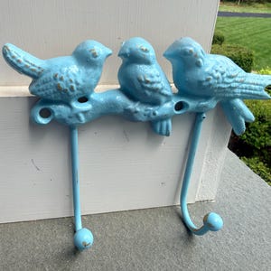 Peut inclure: Un crochet mural en métal bleu clair avec trois oiseaux décoratifs perchés sur une branche. Le crochet comporte deux crochets incurvés et un crochet simple, chacun avec une extrémité arrondie. Les oiseaux ont une finition vieillie.