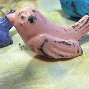 Może przedstawiać: R&oacute;żowa figurka ptaka z wytartym wykończeniem. Ptak ma czarne oko i lekko otwarty dzi&oacute;b. Figurka jest wykonana z metalu i ma rustykalny wygląd.