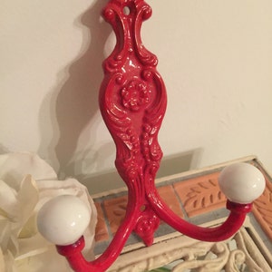 May include: Red metal double hook with white ceramic knobs. The hook has a decorative, ornate design.