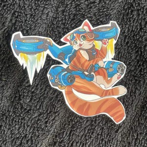 May include: A sticker featuring a cartoon cat riding a blue jetpack. The cat is orange and white with goggles and a brown vest. The jetpack has blue and silver accents with white exhaust trails.