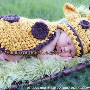 May include: A newborn baby wearing a crocheted giraffe costume, sleeping on a green fur rug. The costume is yellow and brown with a long tail and a hat with ears.
