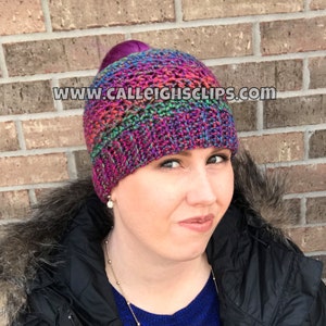 May include: A colorful crochet hat with a hole in the top for a ponytail. The hat is made with a variety of colors, including pink, purple, blue, green, and black. The hat is worn by a person with a dark brown ponytail. The text "www.calleightsclips.com" is visible on the image.