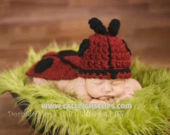 May include: A newborn baby wearing a red and black ladybug crochet hat and matching ladybug costume, sleeping on a bed of green fur.