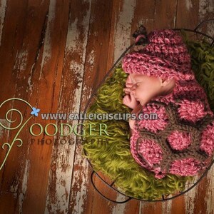Könnte beinhalten: Eine rosa und braune gehäkelte Baby-Mütze mit Blumenmuster. Die Mütze liegt auf einer grün-braunen, flauschigen Oberfläche.