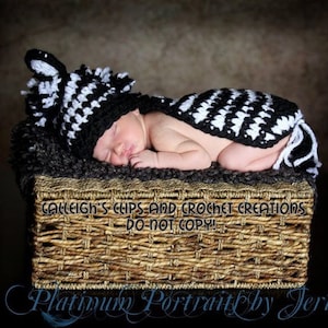 Könnte beinhalten: Ein Neugeborenes trägt eine schwarz-weiße gehäkelte Zebra-Mütze und ein passendes Outfit und liegt in einem Weidenkorb. Das Baby schläft friedlich.