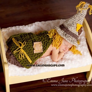 May include: A baby wearing a crocheted scarecrow costume with a yellow straw hat and green and brown fabric. The baby is sleeping on a white and yellow crib.