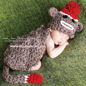 May include: A newborn baby wearing a brown crocheted monkey costume with a red pom-pom on the hat. The baby is sleeping on a green textured surface. The costume has a tail and a face with white details. The text "www.calleightsclips.com" is visible on the costume.