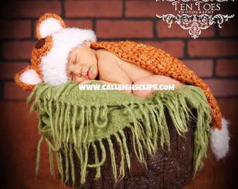 May include: A newborn baby wearing a crocheted fox hat and tail, sleeping in a green and brown basket. The baby is wearing a white hat with brown ears and a brown tail. The basket is made of wood and has a green fabric lining.